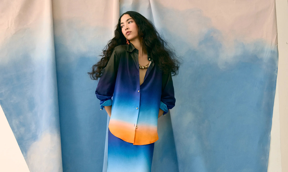 Une mannequin en chemise satinée dégradée du bleu à l'orange, devant la toile peinte en ciel